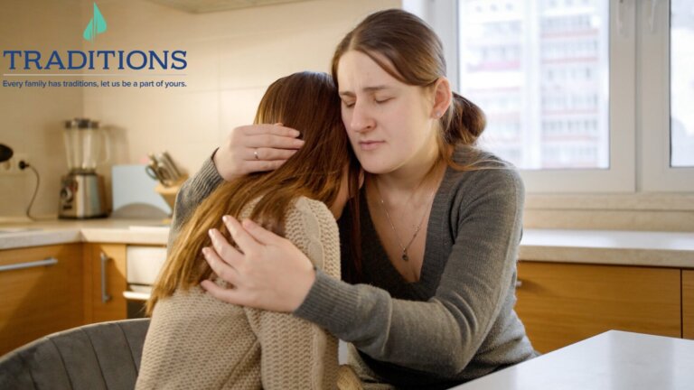 A woman hugging their daughter in a kitchen with a traditions logo on the top left corner