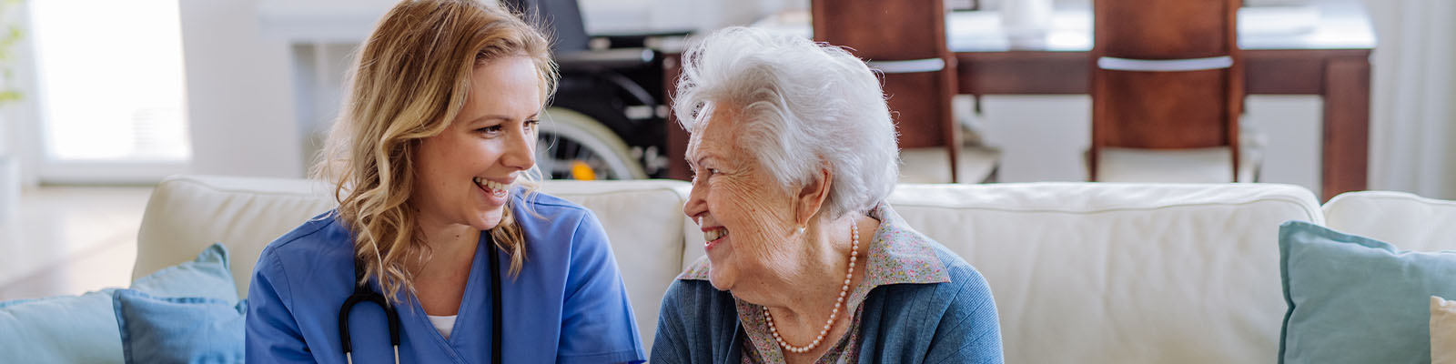 A female senior living resident smiling and sitting on the couch beside her female nursing assistant