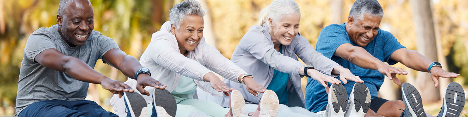 A group of senior men and women sitting outside on the grass together stretching their legs before walking on a walking path